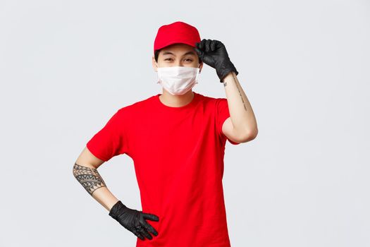 Friendly charismatic asian delivery guy salute client, touch cap and smiling eyes, wear protective gloves and medical mask, contactless delivery concept. Courier in uniform work in carrier company