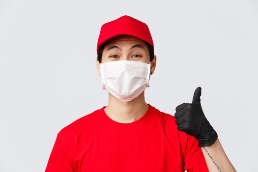 Covid-19, self-quarantine online shopping and delivery concept. Cheerful asian courier in medical mask and protective gloves, wear red uniform, show thumb-up in approval, recommend make safe orders