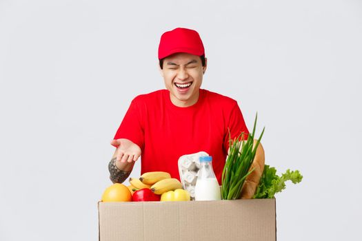 Online shopping, food delivery and internet stores concept. Handsome asian male courier in red uniform, laughing as joking with employees, deliver grocery packages to customer placed order