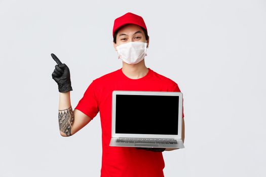 Smiling asian delivery man in uniform, red cap and t-shirt, wearing protective mask and gloves to prevent covid 19 spread, deliver online orders during self-quarantine isolation, invite visit webpage