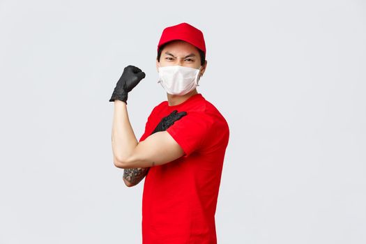 Strong asian guy show-off his strength, carry large parcels at work. Delivery man in uniform flex biceps and grimacing, wear medical mask and gloves, advertisement of carrier and order delivery
