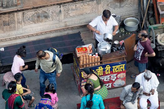 aerial drone shot of road side tea seller in white shirt kurta with small stall preparing tea on a stove and old utensils by putting in milk, leaves, sugar and more as the crowds move around him in the busy city