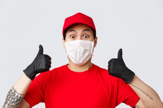 Covid-19, self-quarantine online shopping and delivery concept. Enthusiastic asian courier in red cap and t-shirt uniform, wear medical mask and gloves, thumb-up, recommend safe deliver