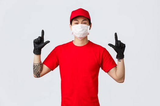 Reluctant or scared, insecure asian guy whining, grimacing as pointing fingers up, wear delivery uniform, red cap and t-shirt, company protect personnel health with protective masks and gloves