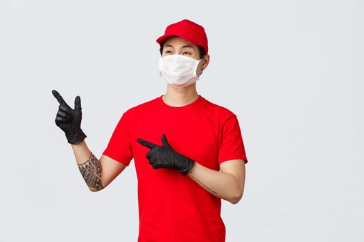 Reluctant and displeased asian delivery guy whining over large order, looking mountain of boxes. Worker of deliver company pointing upper left corner and grimace. Courier in medical mask and gloves
