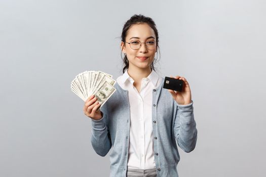 Business, finance and investment concept. Confident good-looking asian girl in glasses, female employee invest money, make good purchase or deposit, show cash dollars and credit card smile satisfied