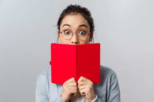 Education, university and people concept. Close-up portrait of surprised and amused young asian girl, widen eyes at amazing interesting story, cover mouth behind red planner, stand grey background