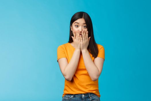 Shocked speechless stunned asian girl stare camera cover mouth palms, raise eyebrows gasping astonished, feel pitty sorry hearing bad news, stand surprised blue background wear yellow t-shirt