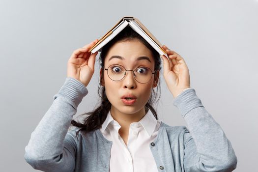 Education, university and people concept. Close-up portrait of wondred and amused asian female in glasses, holding planner, notebook or book on head, realise something interesting, grey background