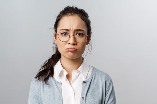 Close-up portrait of gloomy, upset and disappointed young asian female facing failure, losing, regret mjssing interesting event, pouting and sob, frowning camera standing grey background