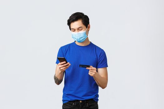 Contactless payment, online shopping during covid-19 and pandemic concept. Young carefree guy place order, insert credit card digits to buy something internet, typing on mobile phone, wear mask