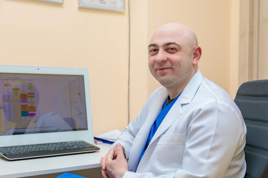 A male ophthalmologist at the workplace at the computer looks at the camera with a smile