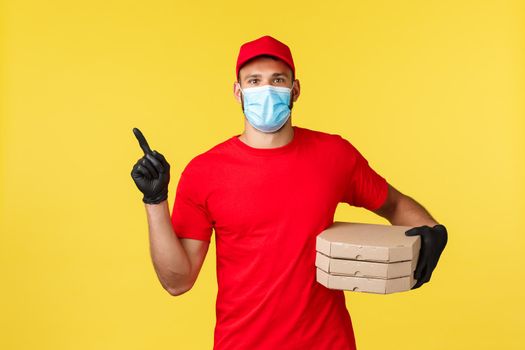 Food delivery, tracking orders, covid-19 and self-quarantine concept. Courier in red uniform and medical mask, provide contactless fast deliver, pointing finger left, holding pizza