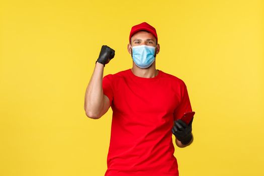 Express delivery during pandemic, covid-19, safe shipping, online shopping concept. Excited courier in medical mask and gloves, using mobile phone and fist pump in success or celebration.