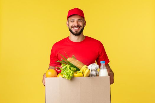 Online delivery, shopping and food order concept. Nice delivery guy, courier in red uniform company, bring grocery order in box to client, smilign friendly, standing yellow background