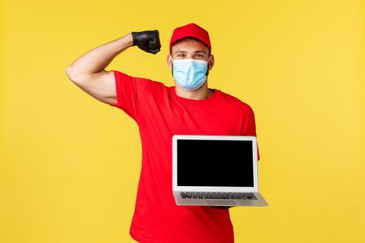 covid-19, safe shopping, express delivery and order tracking concept. Strong handsome courier in red uniform and medical mask, flex biceps and showing laptop screen, smiling