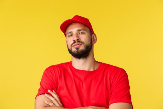 Close-up of confident handsome bearded delivery guy, male courier in red uniform cap and t-shirt, look tough and serious, standing yellow background self-assured, deliver any parcel worldwide