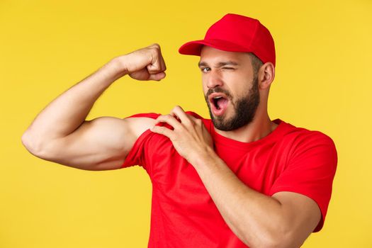 Cheerful handsome delivery guy in red uniform cap and t-shirt, winking at camera as show-off with strong muscles, flex biceps ensure can carry any parcel, courier brag strength over yellow background