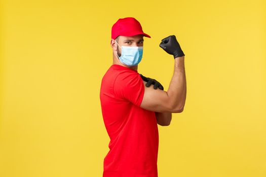 Express delivery during pandemic, covid-19, safe shipping, online shopping concept. Strong young courier in red uniform company and medical mask, show muscles, flex biceps over yellow background