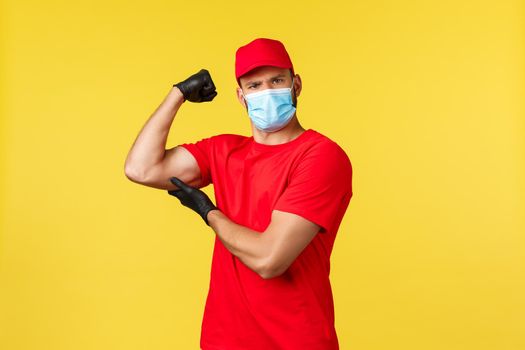 Express delivery during pandemic, covid-19, safe shipping, online shopping concept. Strong and determined courier in red uniform, medical mask and gloves, flex biceps, brag with muscles
