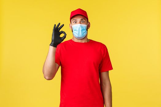 Express delivery during pandemic, covid-19, safe shipping, online shopping concept. Surprised and startled courier showing okay sign, widen eyes impressed, wear red company uniform and medical mask