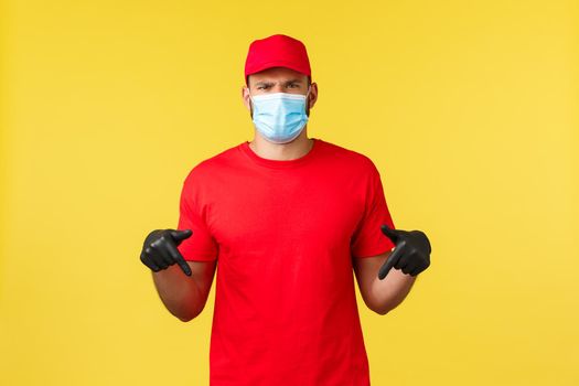 Express delivery during pandemic, covid-19, safe shipping, online shopping concept. Disappointed and upset courier in red uniform, medical mask, pointing fingers down at bad news