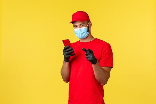 Food delivery, tracking orders, covid-19 and self-quarantine concept. Young courier, employee in medical mask and gloves, using mobile phone and pointing finger camera, yellow background