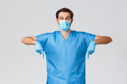 Covid-19, healthcare workers and hospital concept. Cheerful enthusiastic doctor in scrubs, fighting coronavirus, wear protective equipment, medical mask and gloves, pointing down to show promo