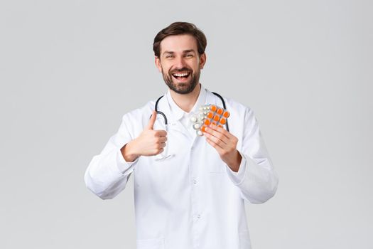 Hospital, healthcare workers, covid-19 treatment concept. Happy cheerful caucasian doctor smiling, show thumb-up in approval and advice medication, vitamins or pills, promote health service