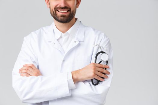 Hospital, healthcare workers, covid-19 treatment concept. Cropped shot of bearded smiling doctor in white scrubs, cross arms like professional physician or therapist, holding stethoscope