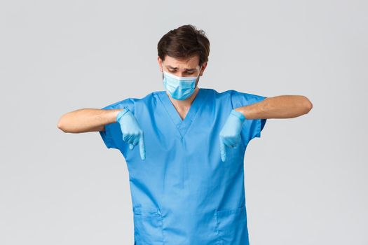 Coronavirus outbreak, healthcare workers fighting disease, hospitals concept. Disappointed doctor showing compassion or sympathy, pointing fingers down, look upset, wear scrubs and gloves