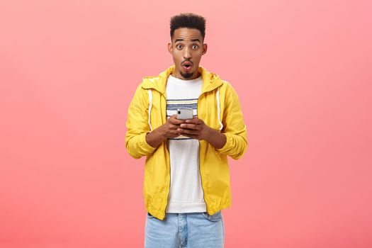 Impressed enthusiastic african american male with curly haircut folding lips in wow sound holding smartphone being excited and thrilled with cool device feautres or new app over pink wall
