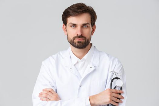 Hospital, healthcare workers, covid-19 treatment concept. Close-up of bearded serious-looking doctor in white scrubs, holding stethoscope, cross arms chest professional pose