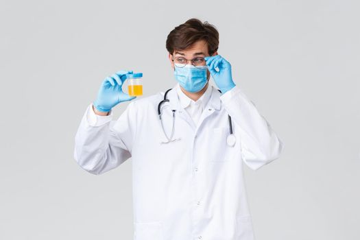 Hospital, healthcare workers, covid-19 treatment concept. Lab technician, doctor examine urine sample in laboratory, wearing protective medical mask and gloves, look intrigued, grey background
