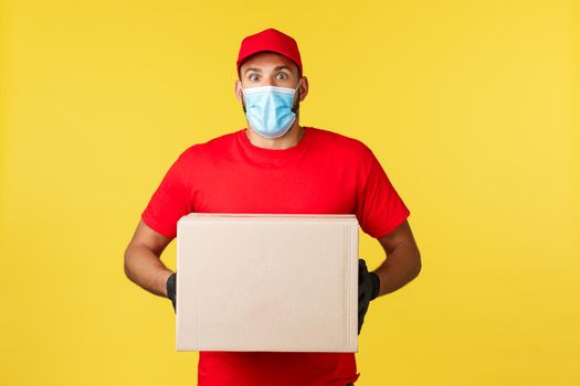 Express delivery during pandemic, covid-19, safe shipping, online shopping concept. Startled and shocked courier in red uniform, medical mask and gloves, holding parcel feel astonished