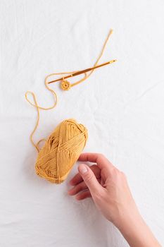Crochet hook and ball of cotton yarn ocher color in a hand on a white table