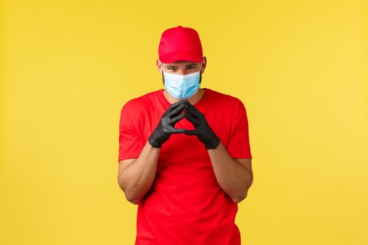 Express delivery during pandemic, covid-19, safe shipping, online shopping concept. Devious and creative young courier in red uniform and medical mask, have plan, scheming over yellow background
