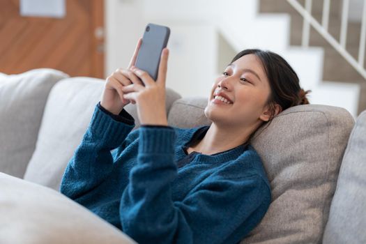 Happy young asian woman lying on sofa at living room and using cell phone at home