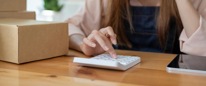 Asian woman entrepreneur using calculator with pencil in her hand, calculating financial expense at home office,online market packing box delivery,Startup successful small business owner,SME, concept
