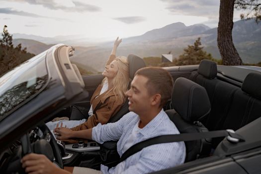 Happy young couple in love driving roofless convertible car holding hands, mountains view