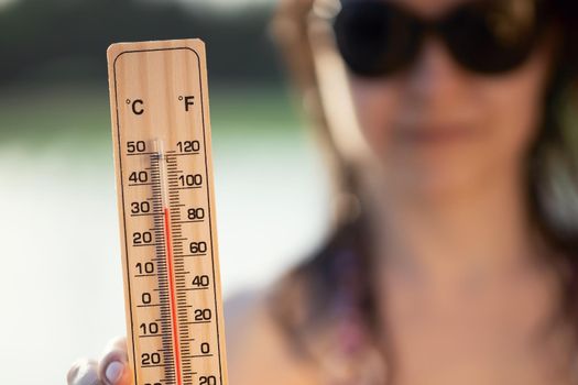 Beautiful young woman in sunglasses with a thermometer in her hand on the background of the lake. Summer vacation and heat concept, selective focus