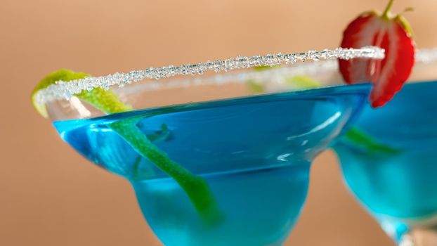 Two glasses with blue margarita cocktail garnished with lime zest and strawberries, selective focus
