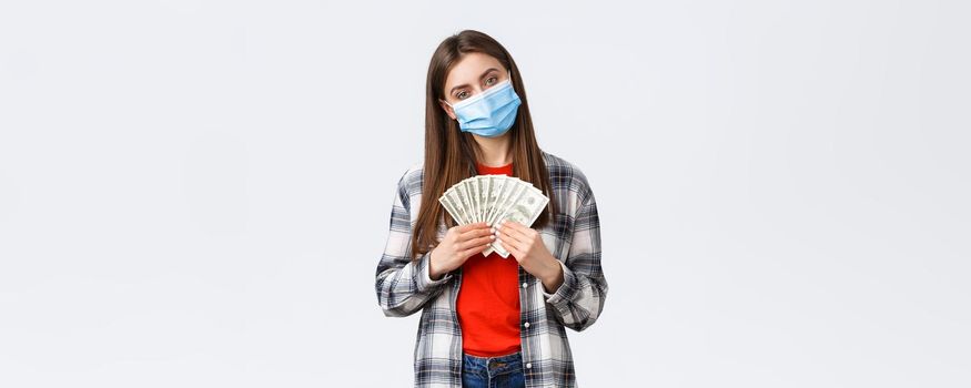 Money transfer, investment, covid-19 pandemic and working from home concept. Pleased young female employee, freelancer in medical mask holding her salary or won cash, white background
