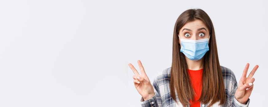 Coronavirus outbreak, leisure on quarantine, social distancing and emotions concept. Close-up of excited and thrilled cute woman in medical mask, showing peace sign or quotes, stare impressed