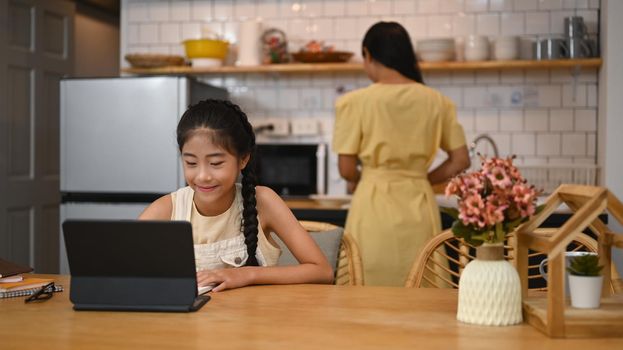 Asian girl having online class on computer tablet, doing homework at kitchen table. Concept of virtual education, homeschooling