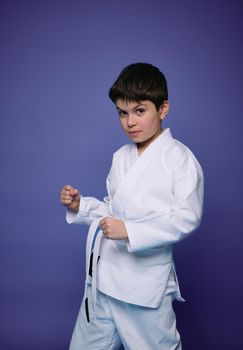 Portrait of a handsome Caucasian teenage boy, aikido wrestler practicing martial skills against purple wall background. Oriental martial arts concept