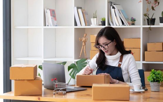 Young beautiful happy asian business woman owner of SME online using laptop receive order from customer with parcel box packaging at her startup home office, online business seller and delivery