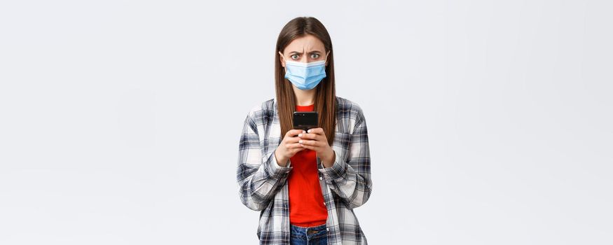 Different emotions, covid-19, social distancing and technology concept. Frustated and confused young woman in medical mask react to strange message, hold mobile phone, frowning camera