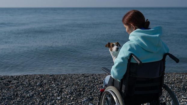 Caucasian woman in a wheelchair cuddling with a dog near the sea.