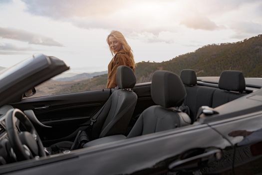 Pretty Caucasian young happy female standing outdoor behind convertible car looking at camera